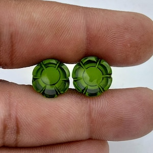 May include: Two round, olive-green gemstones with a carved floral design. The gemstones are held between fingers, showcasing their intricate details and colour. The gemstones are likely for jewellery making.
