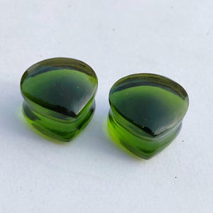 May include: Two green glass teardrop-shaped ear plugs. The plugs have a rounded top and a flared base. The glass is a deep, translucent green color. The plugs are sitting on a white surface.