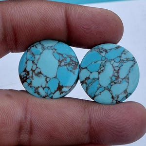 Puede incluir: Dos piedras preciosas redondas de turquesa con un patrón moteado de azul claro y marrón. Las piedras se sostienen entre los dedos, mostrando su superficie pulida y variaciones naturales. Ideal para la fabricación de joyas.