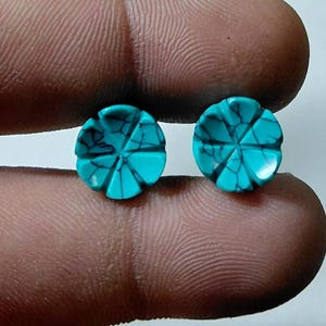 May include: Two turquoise-colored, flower-shaped gemstones. Each gem has a six-petal design with dark veining. The gems are held between fingers, showcasing their intricate carved details and vibrant color. These could be used for jewelry.