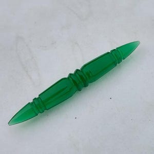 May include: A vibrant green, elongated glass bead with pointed ends and a decorative, ridged center. The bead is a solid, translucent color and appears to be hand-crafted. The object is set against a white background.