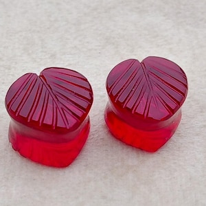 May include: A pair of heart-shaped ear plugs in a deep red hue. Each plug has a leaf-vein pattern carved into the top. The plugs have a translucent quality, with a gradient of red tones.