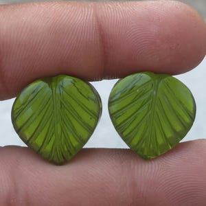 May include: Two green leaf-shaped beads with detailed vein patterns. The beads are held between fingers, showcasing their translucent quality and intricate design. These beads are suitable for jewelry making.