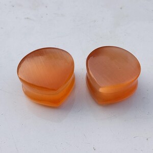 May include: Two orange-colored, teardrop-shaped ear plugs. The plugs have a smooth, polished surface and a slightly translucent appearance. The plugs are designed to fit comfortably in the ear and are made of a durable material.