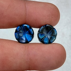 May include: Two round, carved gemstones with a starburst design. The stones are a deep blue color with iridescent flashes of light. The gemstones are held between two fingers.