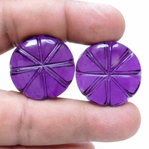 May include: Two round, deep purple gemstones with carved radial patterns. The gemstones are held between fingers against a white background. The carved lines create a starburst effect on each stone, giving them a unique, textured appearance.