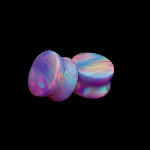 Peut inclure: Une paire d'écarteurs d'oreilles circulaires présentant une finition lisse et irisée avec des teintes tourbillonnantes de violet, bleu, rose et jaune. La surface de ces bijoux de corps reflète la lumière, créant un effet nacré sur les faces.