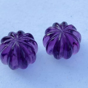 Könnte beinhalten: Zwei geschnitzte, tiefviolette Edelsteinperlen. Jede Perle hat eine blütenartige Form mit gebogenen, geriffelten Blütenblättern. Die Perlen haben eine satte, durchscheinende Farbe und liegen auf einer hellblauen Oberfläche. Diese Perlen könnten für die Schmuckherstellung verwendet werden.