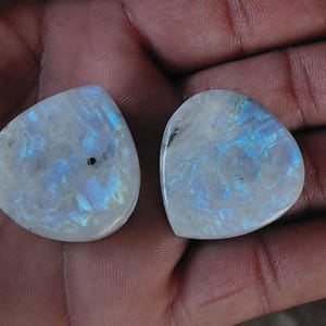 Könnte beinhalten: Zwei polierte, tropfenförmige Mondstein-Cabochons. Die Steine zeigen eine milchig-weiße Grundfarbe mit irisierenden blauen und silbernen Reflexen. Die Cabochons sind glatt und haben einen hohen Glanz, ideal für die Schmuckherstellung.
