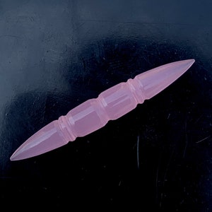 May include: A pink, pointed, glass, decorative object with a textured design. The object is shaped like a long, thin, pointed dagger.