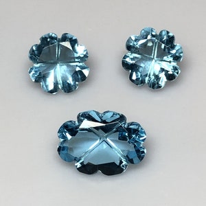 May include: Three blue topaz gemstones cut in the shape of a four-leaf clover. The gemstones are faceted and have a light blue color.