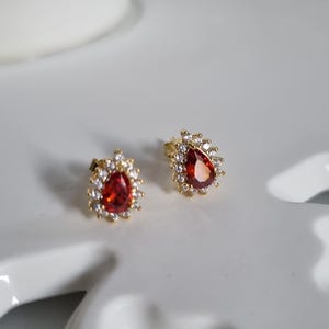 Puede incluir: Un par de pendientes de botón dorados. Cada pendiente presenta una piedra preciosa roja en forma de lágrima, rodeada por un halo de pequeños cristales transparentes. Los pendientes están colocados sobre una superficie blanca decorativa.