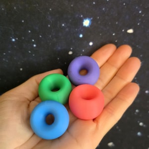 May include: Four small, smooth, torus-shaped objects in solid matte colours: one blue, one green, one purple, and one coral red. Each object features a central circular hole and a rounded, uniform exterior surface, presented as a set of four individual items.