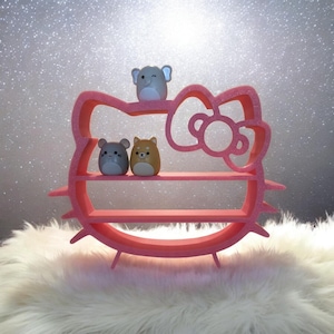 May include: A pink, three-tiered shelf shaped like a bow and face. It displays three small, round figurines: an elephant, a mouse, and a dog. The background is a glittery, silver surface.