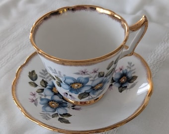 May include: A white teacup and saucer with a gold rim and blue floral pattern. The cup has a handle and the saucer has a scalloped edge.