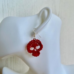 May include: A single red crochet earring shaped like a skull. The earring features a silver-toned metal hook for attachment. The crochet work is detailed with open spaces forming the eye sockets and nasal cavity of the skull design.