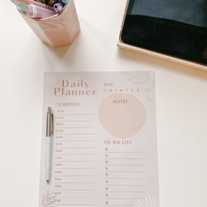 May include: A pink daily planner with a schedule and to-do list. The planner has a light pink background with a darker pink circle and a white pen.