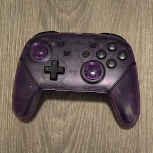 May include: A transparent purple video game controller with black buttons and joysticks. The controller has a plus-shaped directional pad and the letters A, B, X, and Y on the right side.
