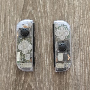May include: Two clear plastic Nintendo Switch Joy-Con controllers with exposed circuit boards and buttons. The controllers are separated and facing forward.