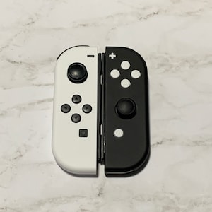 Puede incluir: Un mando de Nintendo Switch blanco y negro con un dise&ntilde;o dividido. El lado izquierdo es blanco con botones negros y el lado derecho es negro con botones blancos.