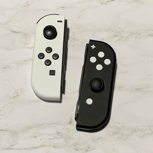 Puede incluir: Dos mandos Joy-Con para Nintendo Switch, uno blanco y otro negro. Ambos mandos tienen la disposici&oacute;n est&aacute;ndar de botones, incluyendo un signo m&aacute;s, un signo menos y cuatro botones direccionales.