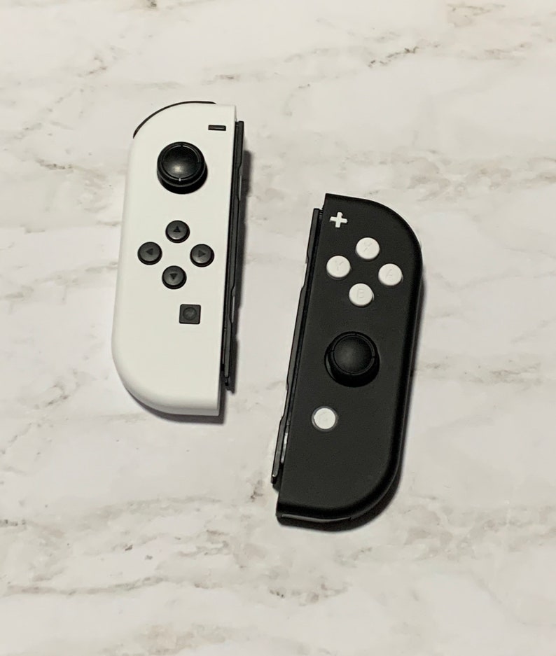 Puede incluir: Dos mandos Joy-Con para Nintendo Switch, uno blanco y otro negro. Ambos mandos tienen la disposici&oacute;n est&aacute;ndar de botones, incluyendo un signo m&aacute;s, un signo menos y cuatro botones direccionales.
