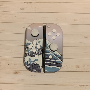 May include: A pair of white and blue Nintendo Switch Joy-Con controllers with a Great Wave off Kanagawa design. The controllers have white buttons and a black center seam.