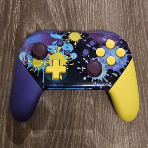 May include: A colorful video game controller with a unique design. The controller features a purple body with yellow and purple accents, including buttons and joysticks. The face of the controller has a splash art design with blue, yellow, and white paint splatters.