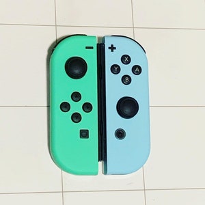May include: A pair of teal and light blue Nintendo Switch Joy-Con controllers. The left Joy-Con is teal and the right Joy-Con is light blue. The controllers have black buttons and a black analog stick.