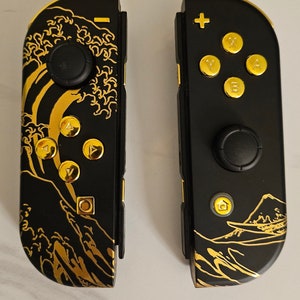 May include: A pair of black and gold Nintendo Switch Joy-Con controllers with a wave and mountain design. The left Joy-Con has a gold wave design and the right Joy-Con has a gold mountain design.