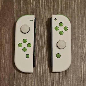 May include: A pair of white Nintendo Switch Joy-Con controllers with green buttons. The controllers have a black stripe down the middle and a white button on the top.