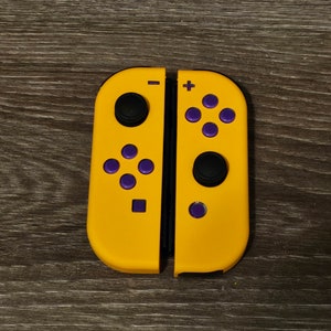 May include: A pair of yellow Joy-Con controllers with purple buttons and black joysticks. The controllers are split, showing the individual components. The design is symmetrical, with a plus and minus button on each side.