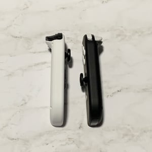 Puede incluir: Dos mandos Joy-Con para Nintendo Switch, uno blanco y otro negro, se muestran uno al lado del otro sobre una superficie de m&aacute;rmol blanco.