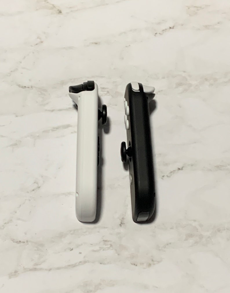 Puede incluir: Dos mandos Joy-Con para Nintendo Switch, uno blanco y otro negro, se muestran uno al lado del otro sobre una superficie de m&aacute;rmol blanco.