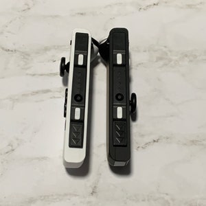 Puede incluir: Dos mandos Nintendo Switch Joy-Con, uno negro y otro blanco, se muestran uno al lado del otro. Ambos mandos tienen la misma disposici&oacute;n de botones, incluyendo un pad direccional, cuatro botones frontales y dos botones de hombro.