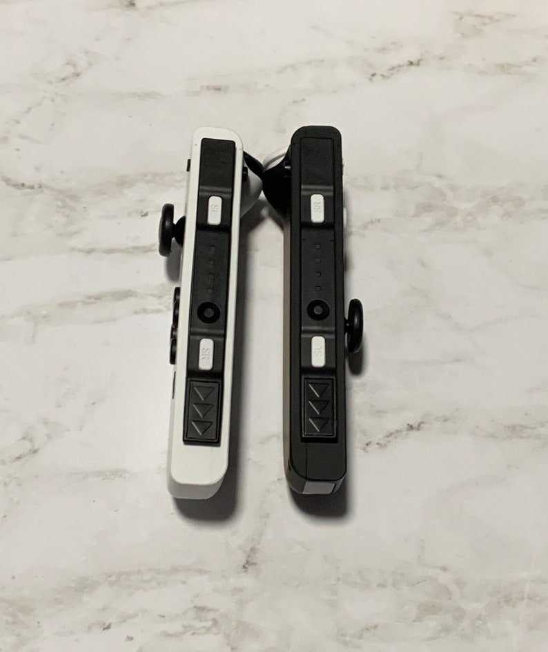 Puede incluir: Dos mandos Nintendo Switch Joy-Con, uno negro y otro blanco, se muestran uno al lado del otro. Ambos mandos tienen la misma disposici&oacute;n de botones, incluyendo un pad direccional, cuatro botones frontales y dos botones de hombro.