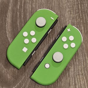 May include: A pair of green Nintendo Switch Joy-Con controllers with white buttons. The controllers are separated and show the front and back sides.