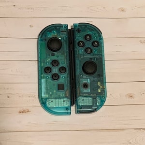 May include: A pair of transparent blue Nintendo Switch Joy-Con controllers with black buttons. The controllers are separated and show the front and back sides.