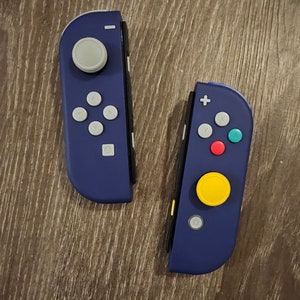 May include: Two purple Nintendo Switch controllers. The left controller has a gray joystick and a standard button layout. The right controller has a yellow button, a red button, and a teal button.