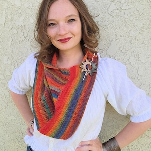 May include: A woman wearing a white shirt with a crocheted scarf in shades of red, orange, yellow, green, and blue. The scarf is worn around the neck and has a decorative pin in the shape of a sun.