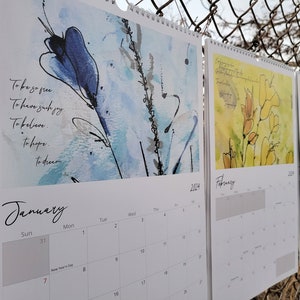 Three calendars hanging in a row on a chain link fence showing the months of January, February, and March which feature blue and yellow loose floral paintings and poetry.