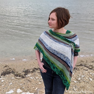 May include: A woman wearing a green, blue, and white striped crochet poncho with a silver pin. She is standing on a beach with water in the background.