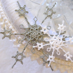 以下が含まれることがあります： 銀色と白の手作りの、かぎ針編みの雪の結晶のクローズアップ。 雪の結晶は、白いレースのトリムが付いた白いテクスチャのある表面に配置されています。 雪の結晶は繊細で複雑なデザインです。