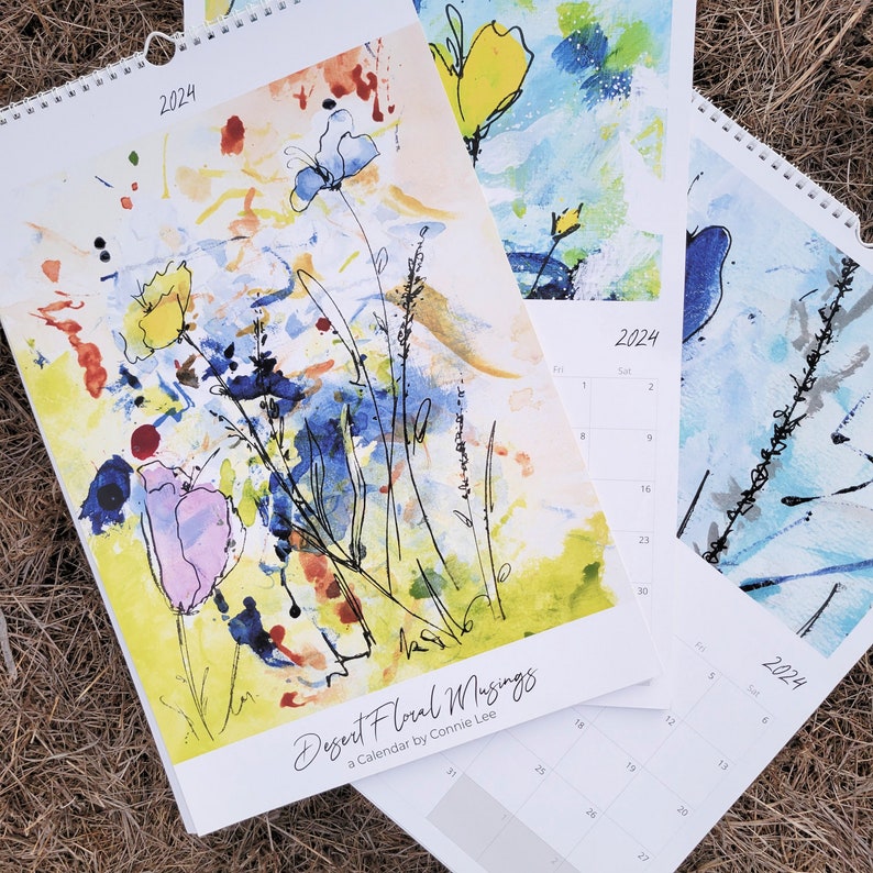 Three 2024 Desert Floral Musings wall calendars laid out on the dried grass with two of them open to different months behind the first calendar which is showing the full painting on the front cover.