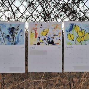Three 2024 wall calendars hanging on a chain link fence showcasing the abstract floral images for the months of July, August, and September. They are primarily blue and yellow.