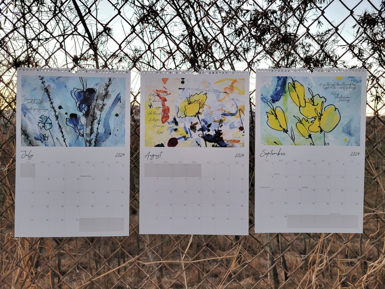 Three 2024 wall calendars hanging on a chain link fence showcasing the abstract floral images for the months of July, August, and September. They are primarily blue and yellow.