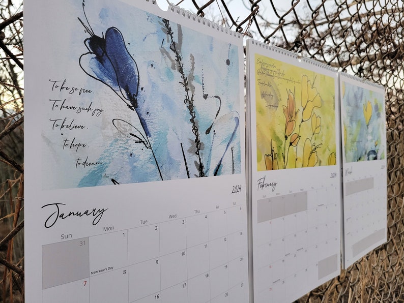 Three calendars hanging in a row on a chain link fence showing the months of January, February, and March which feature blue and yellow loose floral paintings and poetry.
