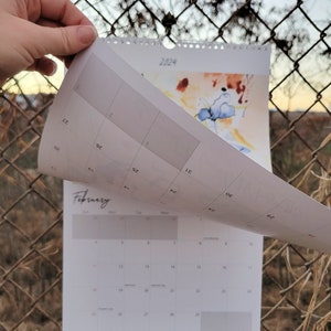 A hand turning the first page of a 2024 Desert Floral Musings Calendar that is hanging up on a chain link fence with the hint of a sunset in the background.