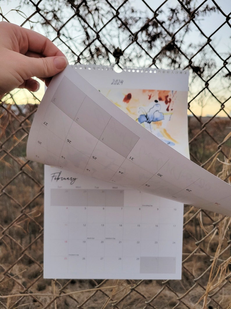 A hand turning the first page of a 2024 Desert Floral Musings Calendar that is hanging up on a chain link fence with the hint of a sunset in the background.
