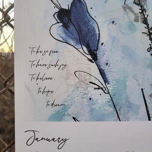A close up image of the first page of the 2024 Desert Floral Musings wall calendar showing an abstract blue flower for the month of January as well as a few lines of poetry, which reads: To be so free. To have such joy. To believe. To hope. To dream.